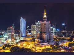 Nighttime cityscape of Warsaw featuring the illuminated Palace of Culture and Science surrounded by modern skyscrapers and office buildings.
