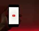 A hand holding a smartphone with the YouTube logo lit up on the display against a red background