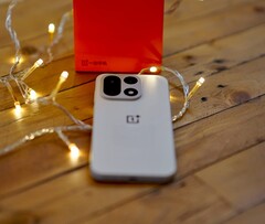 A OnePlus 15 lies on a wooden table surrounded by warm string lights, with a bright red OnePlus retail box positioned in the background.