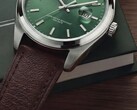 Close-up of the Timex Legacy Day and Date watch, showing its green sunray dial, silver-tone bezel and brushed lugs, ridged crown, and textured brown leather strap against a dark green box and wooden surface.